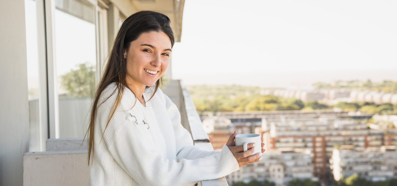 portrait-smiling-young-woman-standing-balcony-holding-white-coffee-cup